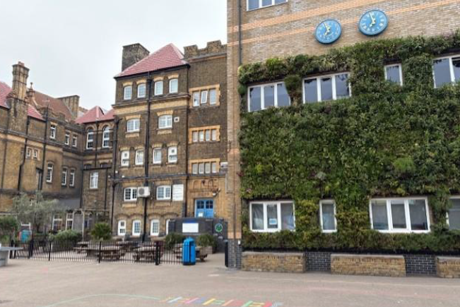 Photograph of a school courtyard featuring a brick building with a green wall covered in plants and two blue clocks mounted above it. 