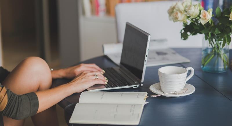 We can see the arms, hands and knee of a person sitting at a laptop at a table. There's a notebook open on the table, also a white cup and saucer, and a vase of flowers. 