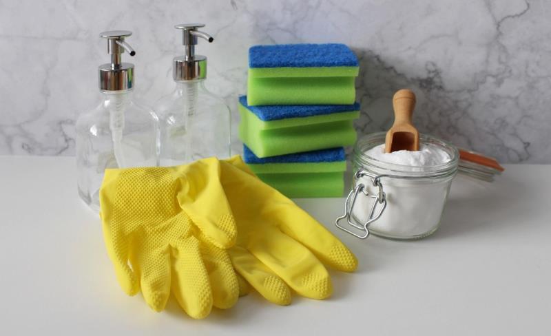 Photograph of cleaning supplies arranged on a white surface with a marble background, including two clear pump bottles, a stack of green and blue sponges, a glass jar filled with white powder and a wooden scoop, and a pair of yellow rubber gloves. 