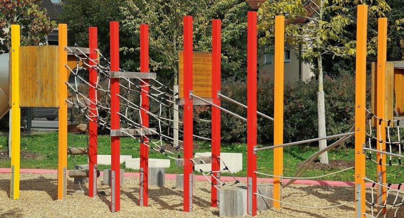 Photograph of an outdoor playground structure featuring vertical poles in yellow, orange, and red colours connected by rope bridges and climbing nets. The setup includes wooden platforms and a woodchip ground surface, designed for children's physical activity and play.