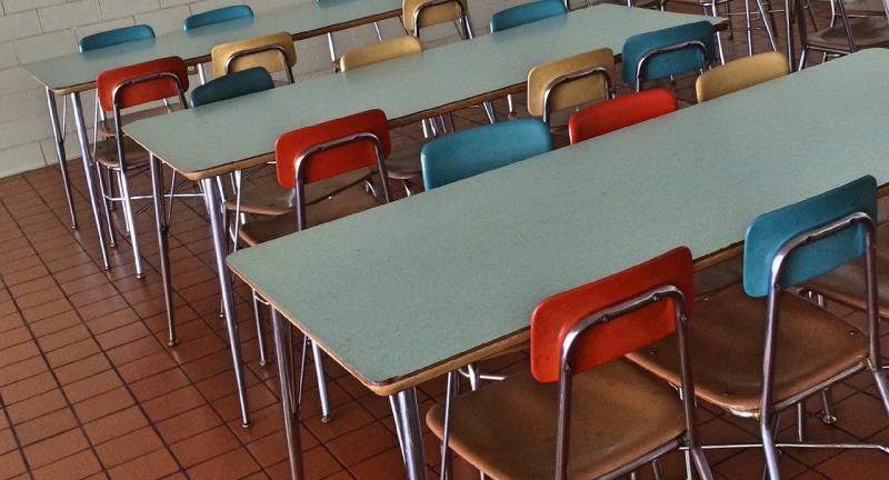 Photograph of a cafeteria or dining area featuring multiple rectangular tables with metal legs and laminate tops. Chairs around tables have metal frames and seats in various colors, including red, blue, brown, and beige, arranged in rows on a tiled floor.