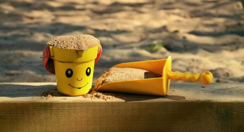 Photograph of a yellow toy bucket with a smiling face and red handles filled with sand, placed on a wooden surface next to a yellow plastic shovel also filled with sand. Background shows a sandy play area with soft sunlight and shadows, suggesting a playful outdoor setting.