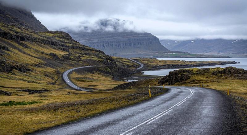 Photograph of a winding road along a rugged coastline with grassy hills and mountains partially covered by clouds in the background. The wet asphalt road features white lane markings and yellow roadside markers, highlighting a remote and scenic landscape under overcast skies. We can see a small white car in the distance. 