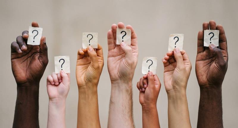 Photograph showing seven raised hands with diverse skin tones, each holding a white card marked with a question mark. Composition symbolizes shared uncertainty, inquiry, or unanswered questions across different people.