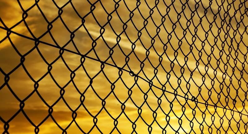 A black chain link fence in close up, against a golden yellow sky. 